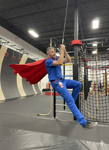 LNA coach in a Superman costume swinging on the ninja course